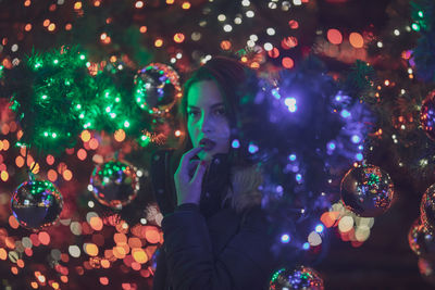 Thoughtful young woman standing amidst illuminated christmas tree at night
