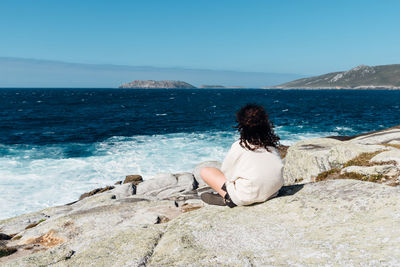 Rear view of woman sitting on rock by sea against sky