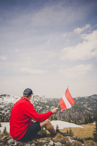 Side view of man holding austrian flag while sitting on mountain against sky during winter
