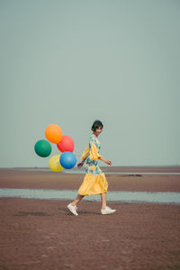 Full length portrait of young woman holding colorful balloons at beach