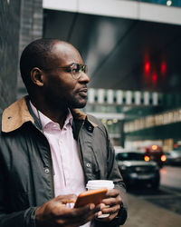 Businessman holding coffee and mobile phone looking away while standing on road in city