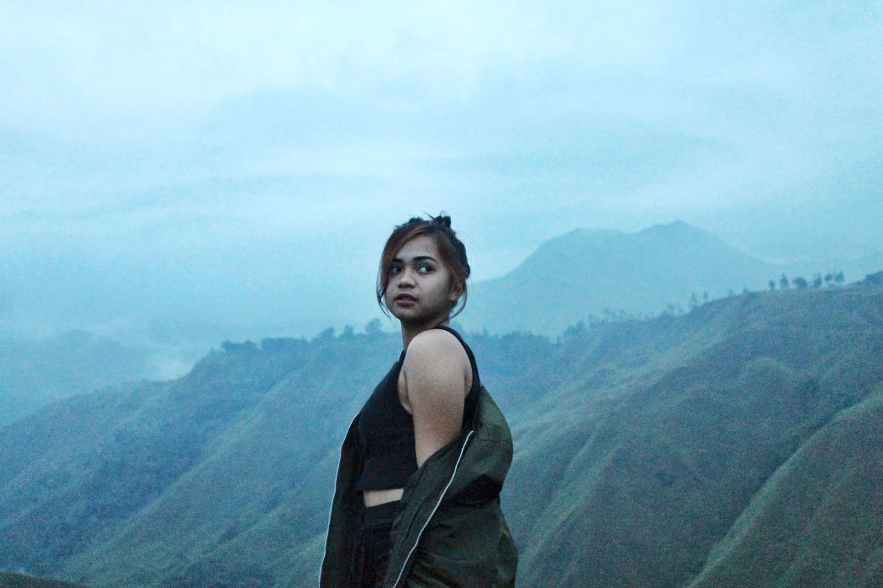 mountain, one person, scenics - nature, leisure activity, sky, young adult, beauty in nature, real people, lifestyles, mountain range, tranquility, nature, standing, environment, tranquil scene, non-urban scene, day, young women, casual clothing, outdoors, contemplation, looking at view, hairstyle