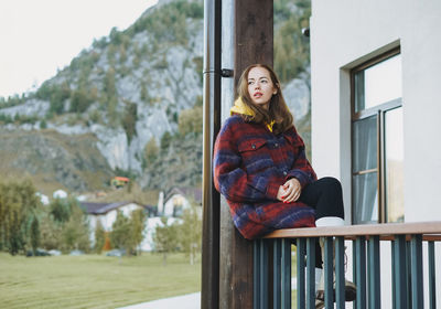 Young woman in coat sitting on wooden fence near cottage on mountains background