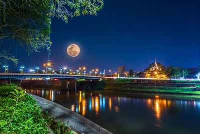 Illuminated bridge over river with temple against sky at night