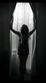Full length of woman standing amidst curtains against window at home