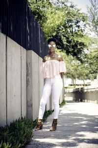 Young woman wearing sunglasses standing on footpath by concrete fence