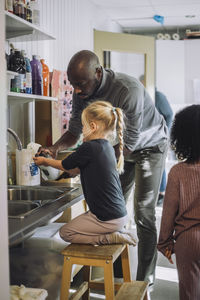 Male teacher giving liquid soap to girl kneeling on stool near sink at kindergarten