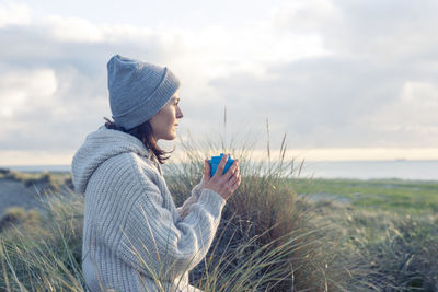 Woman outside in knitwear enjoying a hot drink while looking out to sea.