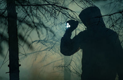 Silhouette man holding flashlight standing by tree in forest at night