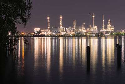 Illuminated factory by river against sky at night