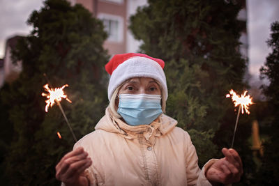 Portrait of senior woman wearing mask holding sparklers against trees