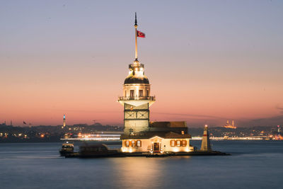 Illuminated building by lighthouse in city against sky during sunset