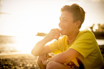 Close-up of teenage boy smiling while sitting at beach against sky during sunset