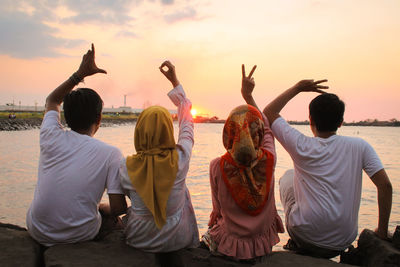 Rear view of friends gesturing while sitting on rocks against sea during sunset