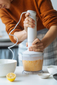 Crop cook mixing pumpkin and checking bowl holding blender in hand on table with citrus in light kitchen