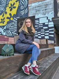 A young woman looks directly into the camera against the backdrop of a painted wall in the fall