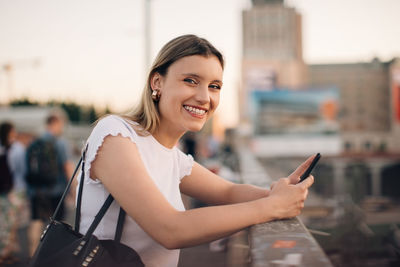 Portrait of smiling young woman holding mobile phone while leaning on railing at bridge in city