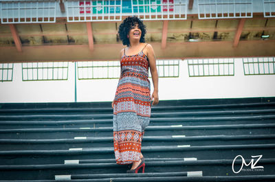 Full length of smiling young woman standing against staircase