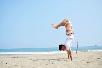 Side view of man doing handstand at beach against clear blue sky during sunny day
