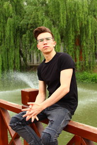 Portrait of young man sitting against trees on railing