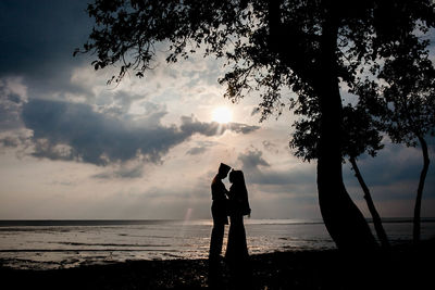 Silhouette couple standing at beach against sky during sunset