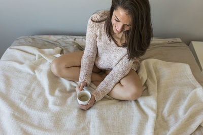 High angle view of woman holding coffee cup while sitting on bed