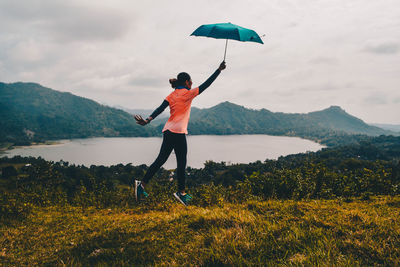 Rear view of woman with umbrella jumping on land near lake against cloudy sky 