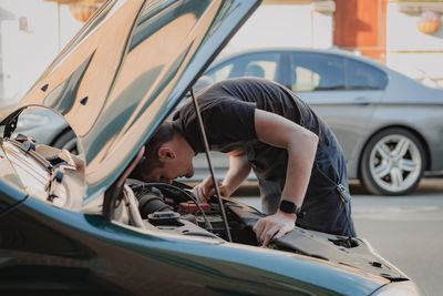 A young man looks into the open hood of a car.
