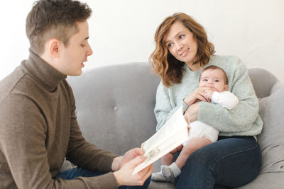 Father reading book while mother sitting with baby at home