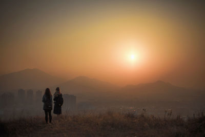 Rear view of friends standing on mountain peak by city against sky during sunset