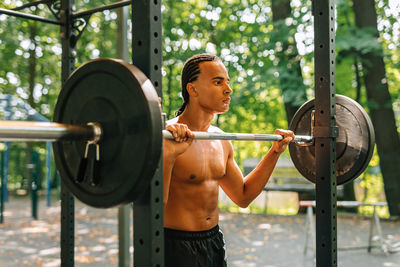 Shirtless man holding barbell while standing outdoors