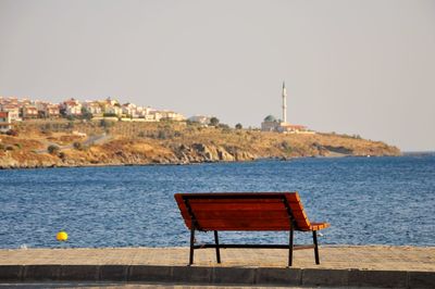 Empty bench by sea against buildings against clear sky