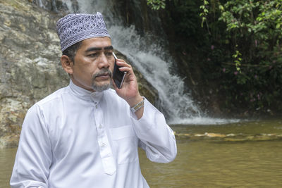 Thoughtful man talking on phone while sitting against waterfall