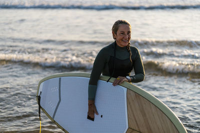 Smiling woman holding paddleboard standing in sea at dawn