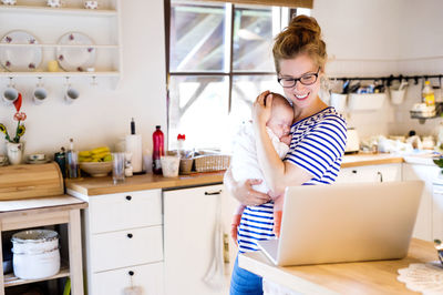 Happy mother with baby in kitchen looking at laptop