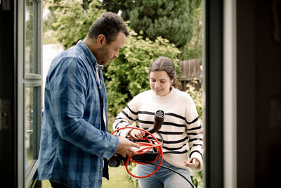 Man and woman talking to each other while holding electric car charger seen through doorway