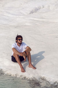 Man with a beard in a white t-shirt and shorts sitting on a white mountain in pamukkale