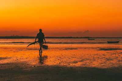 Silhouette of a fisherman walking at beach against sky during sunset