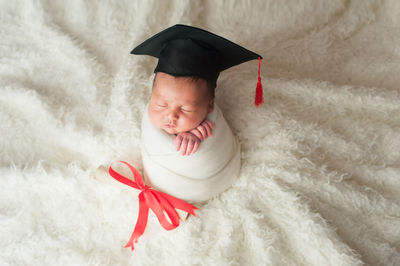 High angle view of cute baby boy wrapped in blanket wearing mortarboard sleeping on bed