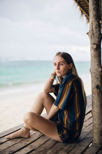 Young woman sitting on shore at beach against sky