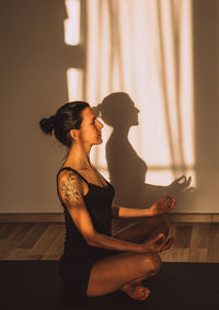 W
woman practicing yoga meditation on sport mat in a room with sunset or sunrise light with shadows.