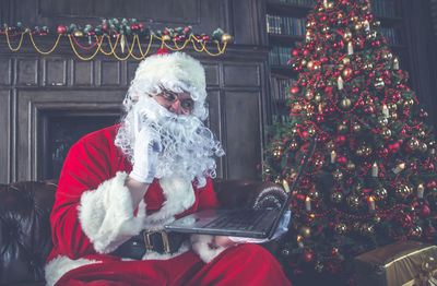Man wearing santa claus costume using laptop while sitting by christmas tree on sofa at home