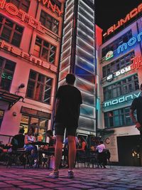 Rear view of people walking on illuminated street amidst buildings at night