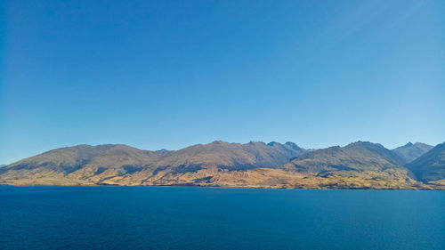 Scenic view of sea by mountains against clear blue sky
