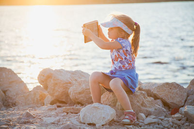 Full length of girl holding digital tablet sitting on rock at beach