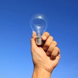 Close-up of hand holding light bulb against blue sky