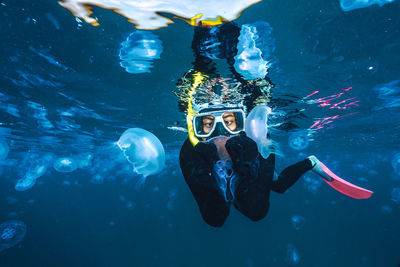 A woman in a black wetsuit is in the water with jellyfish. the jellyfish are floating around her