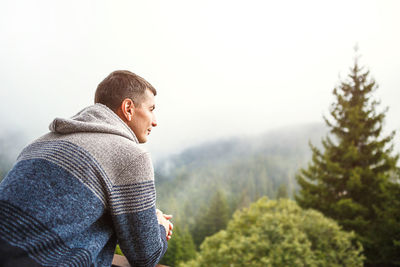 A young man in the morning looks from the hotel balcony with a view of the mountains, the fog