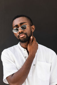 Confident african american male wearing stylish sunglasses and white t shirt standing on street while looking at camera and touching neck