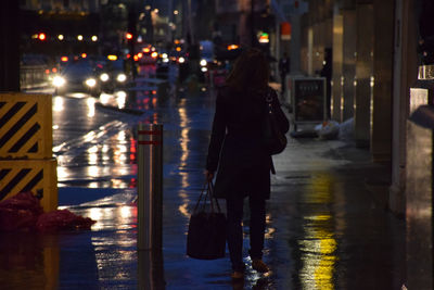 Rear view of woman walking on wet sidewalk in city at night